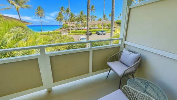 A balcony with a view of the ocean and palm trees, a wicker chair, and a small table. - Ocean view A/C, pool/hot tub coconut marketplace (Kapaʻa)