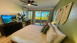 A bedroom with a bed, a television, a desk, and a view of the beach through large windows. - Ocean view A/C, pool/hot tub coconut marketplace (Kapaʻa)
