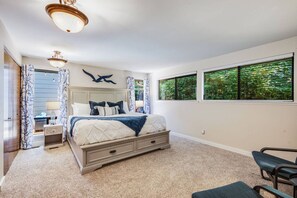 Un dormitorio con cama, mesitas de noche, cómoda, silla y ventanas amplias con persianas.