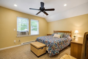 A bedroom with a bed, a ceiling fan, a window with blinds, a bedside table with a lamp, and a small wicker basket.