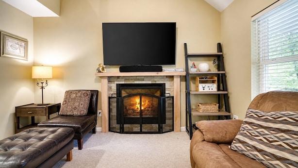 A living room with a fireplace, a flat-screen TV, a sofa, a bench, and a lamp. - Serenity Cottage Deck, Game Room & Fire Pit near Narrowsburg Masthope (Lackawaxen)