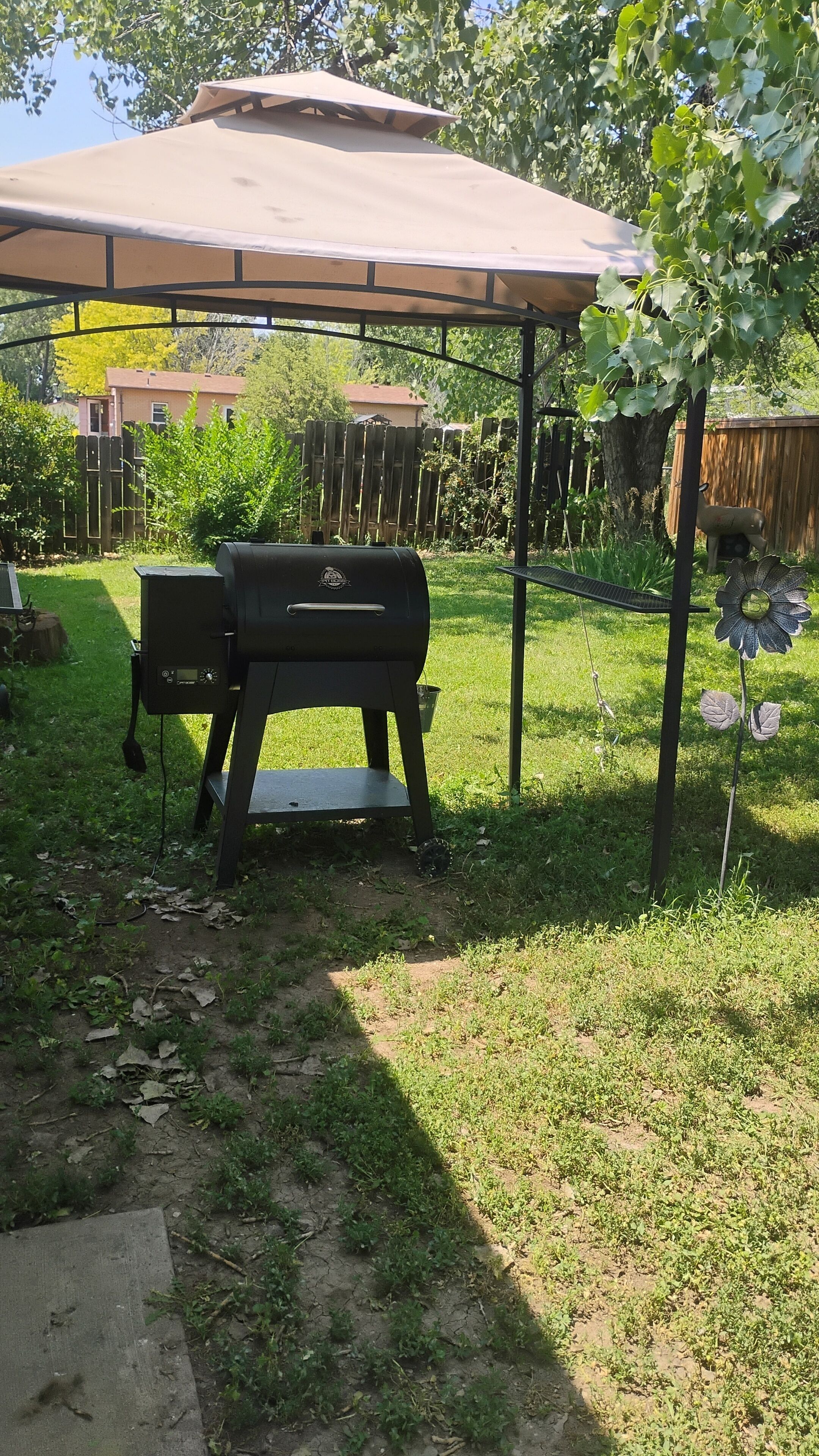 Ein Garten mit Grill unter einer überdachten Terrasse, umgeben von einem Holzzaun und Grünpflanzen.