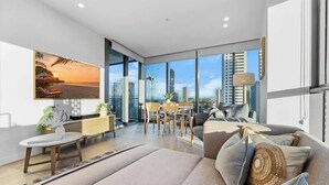 A modern living room with a large window, a sofa, a dining table, and a view of the cityscape. - NEW Ocean view 1BR | Pool, Parking & Walk to Beach (Broadbeach)
