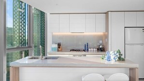 A modern kitchen with a large island, white cabinetry, and a view of the city through large windows. - NEW Ocean view 1BR | Pool, Parking & Walk to Beach (Broadbeach)