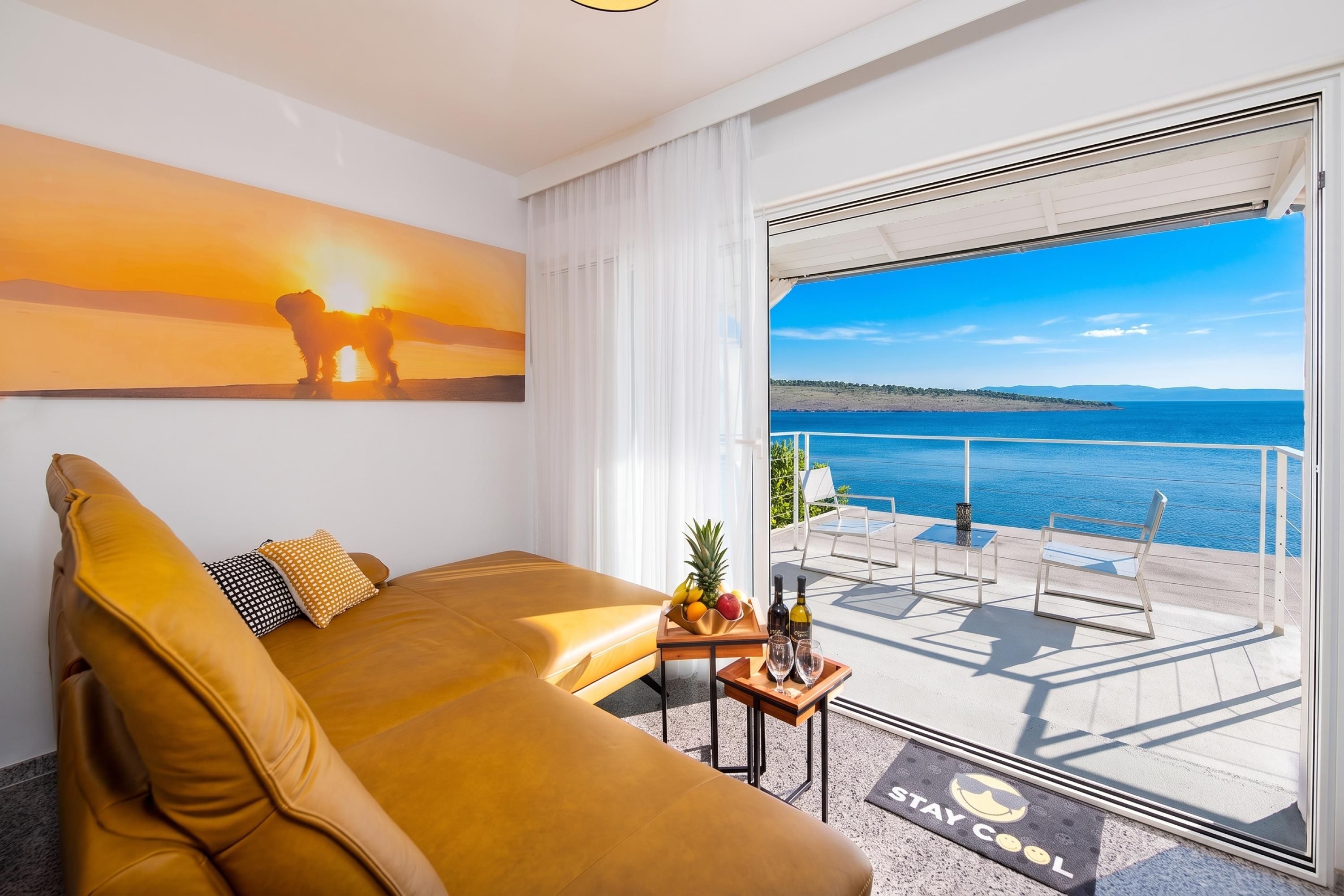 A modern hotel room with a large sofa, a painting of a sunset, a balcony with a view of the sea, and a small table with fruit and wine.