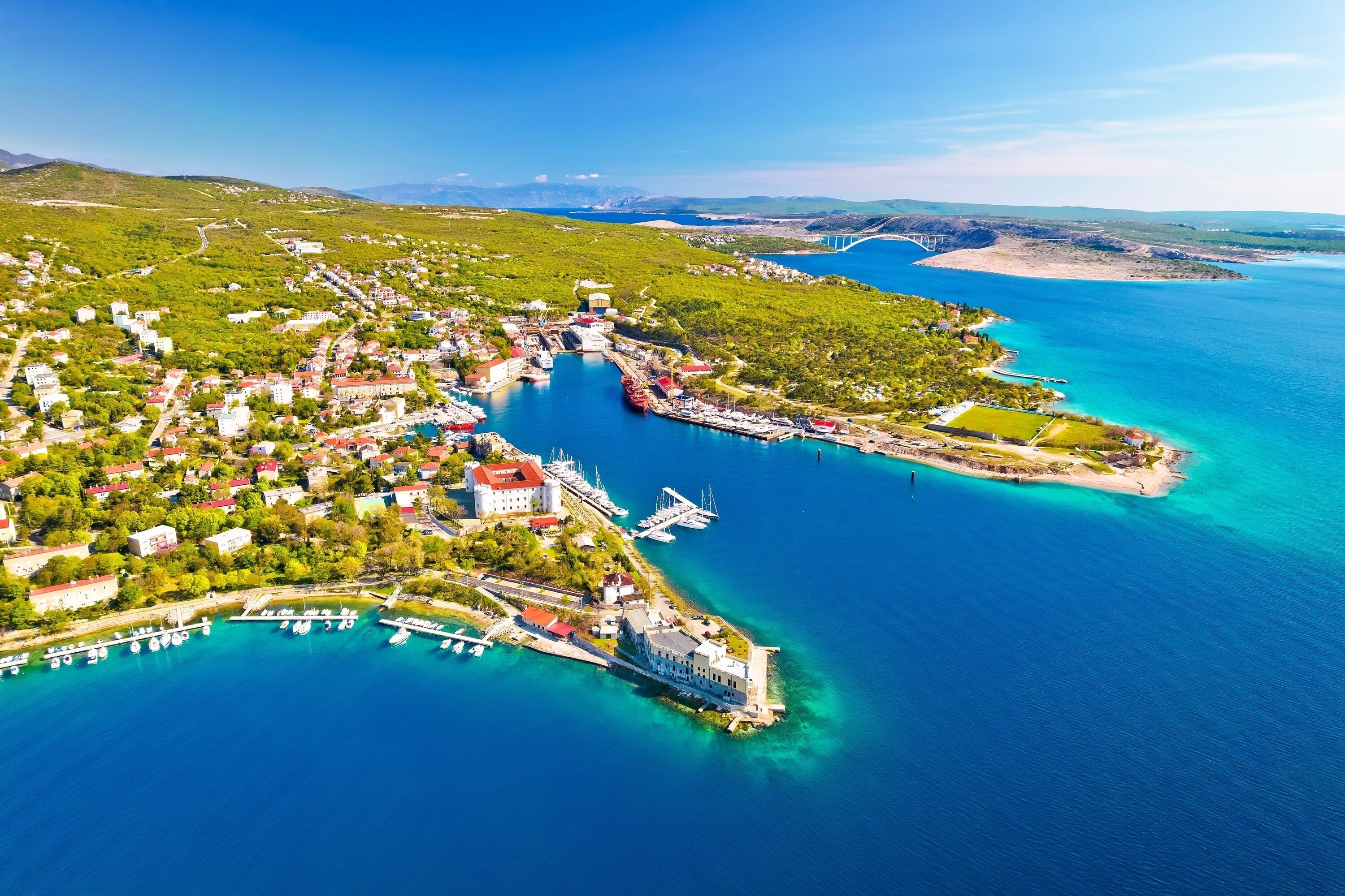 An aerial view of a coastal town with a marina, buildings, and a bridge in the background.