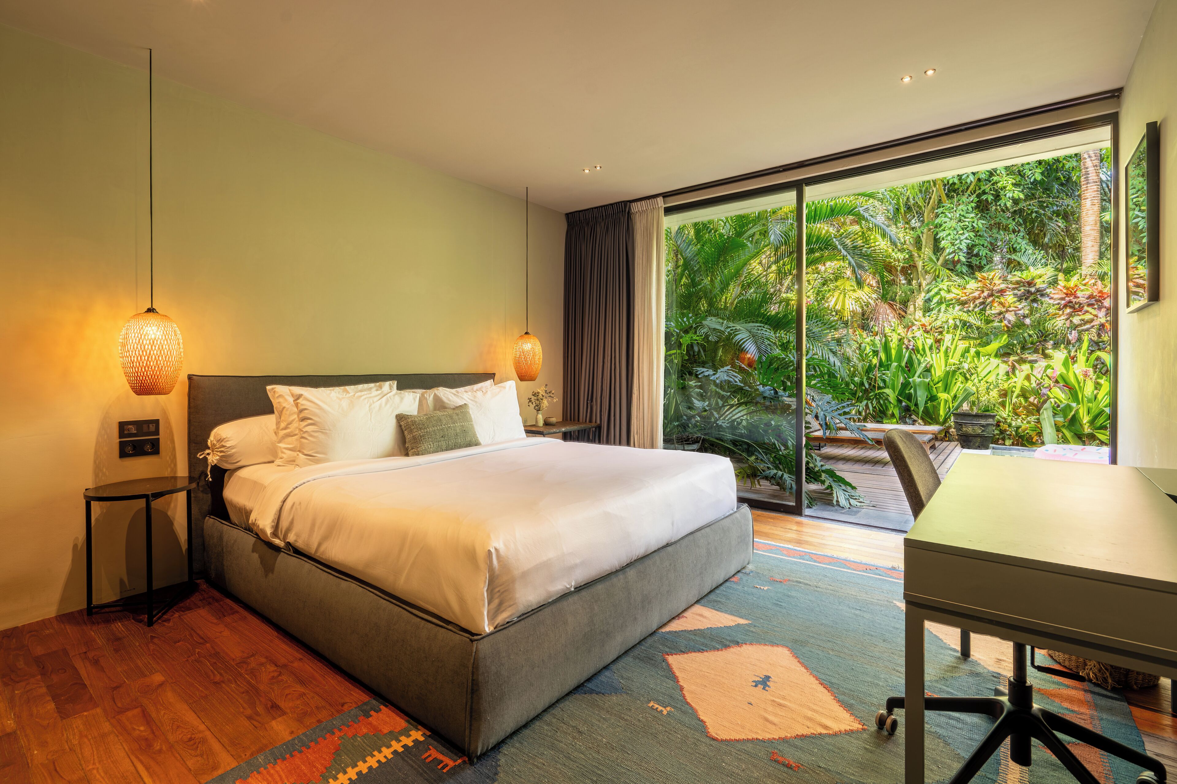 A bedroom with a large bed, a desk, and a view of a lush garden through a sliding glass door.