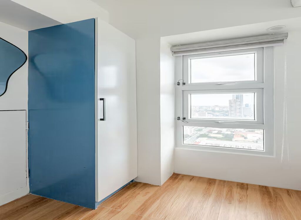 A room with a blue wardrobe, a window with blinds, and a view of a cityscape.