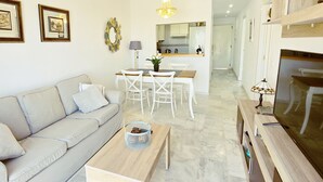 A modern living room with a sofa, a dining table with chairs, a coffee table, a chandelier, and a kitchen area in the background.
