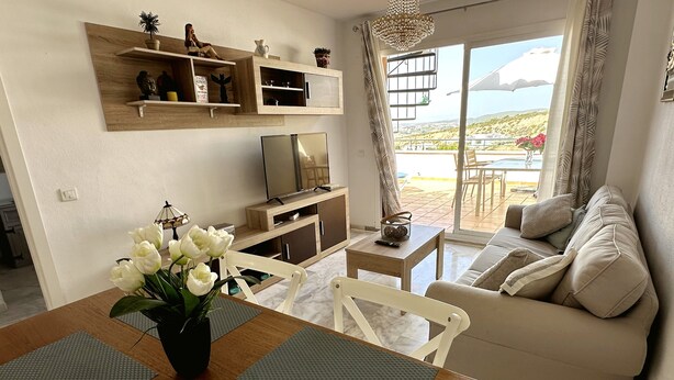 A modern living room with a wooden dining table, white chairs, a beige sofa, and a TV on a wooden stand.