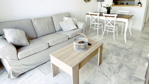 A modern living room with a grey sofa, a wooden coffee table, and a dining area with white chairs and a table.