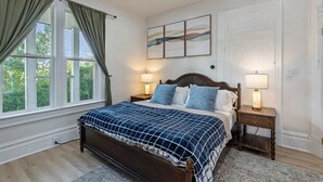 A bedroom with a large bed, two bedside tables with lamps, a window with curtains, and a painting on the wall.
