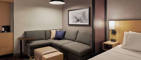 A modern hotel room with a grey sectional sofa, a wooden bench, a small bedside table, a wall-mounted lamp, and a framed artwork.