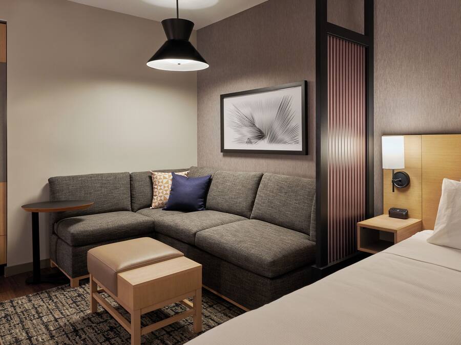 Una habitación de hotel moderna con un sofá seccional gris, un banco de madera, una mesita de noche pequeña, una lámpara de pared y una obra de arte enmarcada.