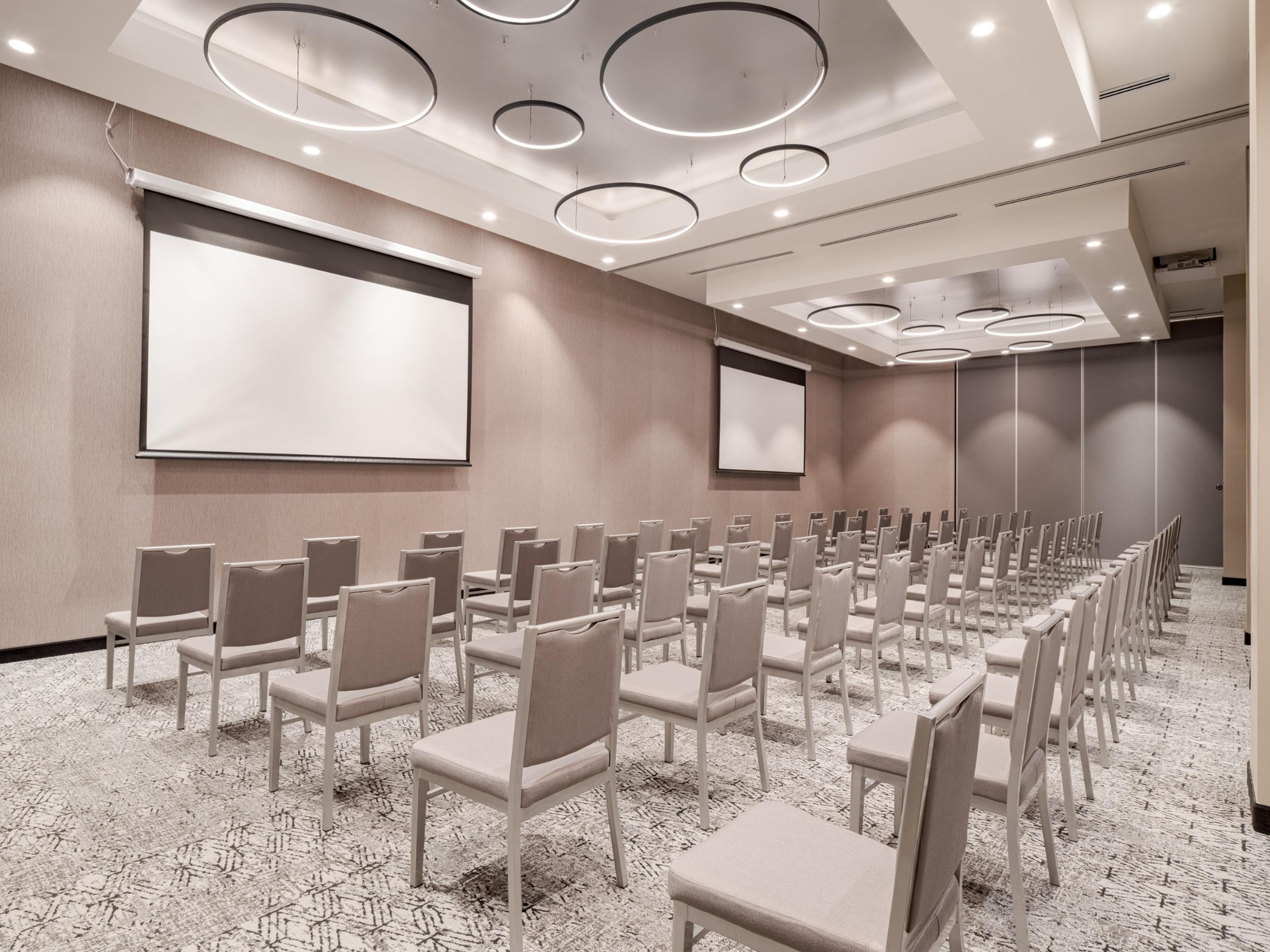 Un salón de conferencias con filas de sillas, una pantalla grande y luces circulares en el techo.