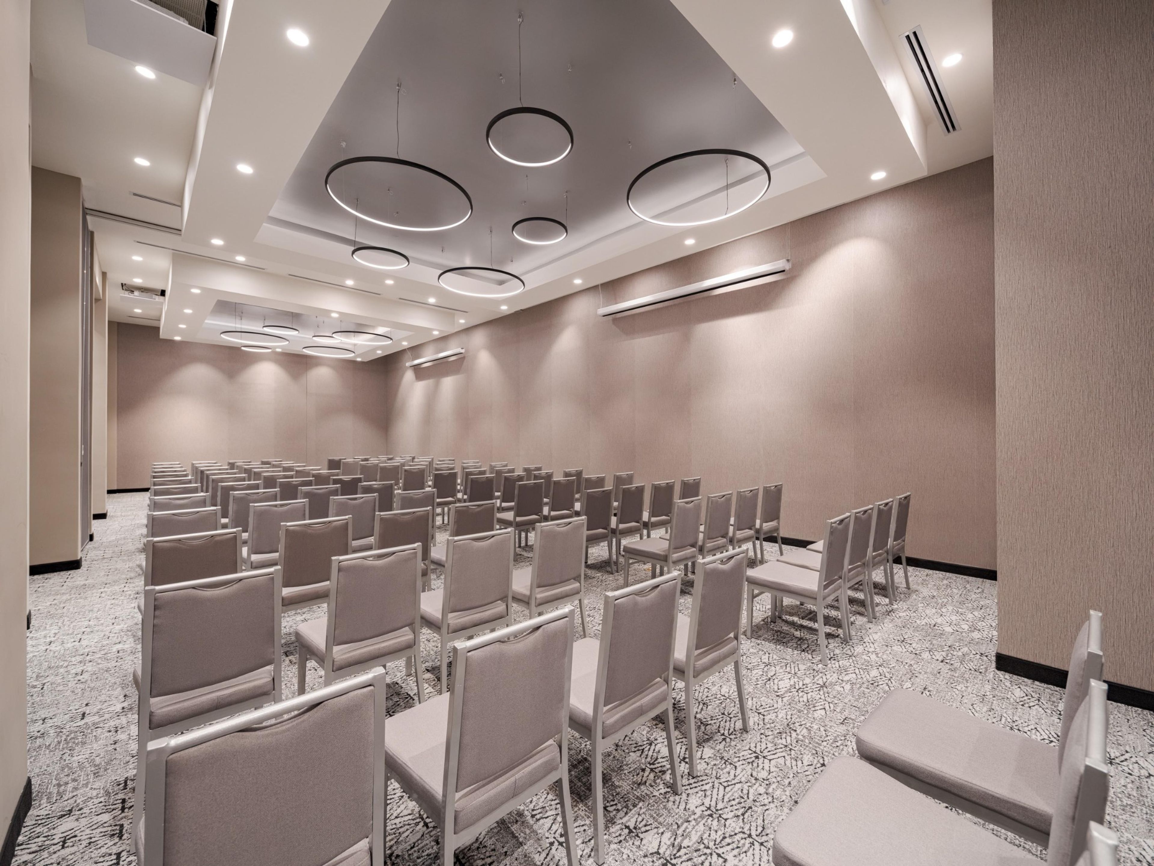 Un salón de conferencias con filas de sillas, un techo con luminarias circulares y suelo cubierto de alfombra.