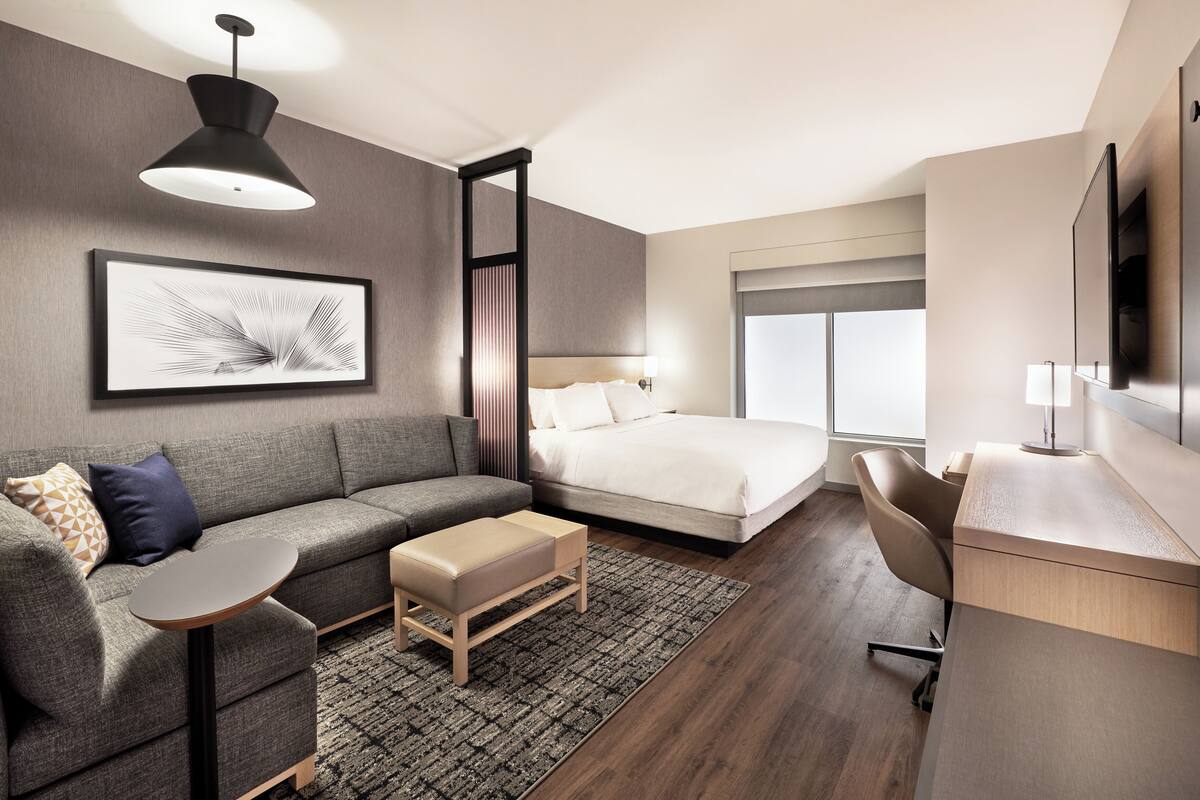 A modern hotel room with a grey sofa, a bed with white linens, a desk with a chair, a large window, and a mounted TV.