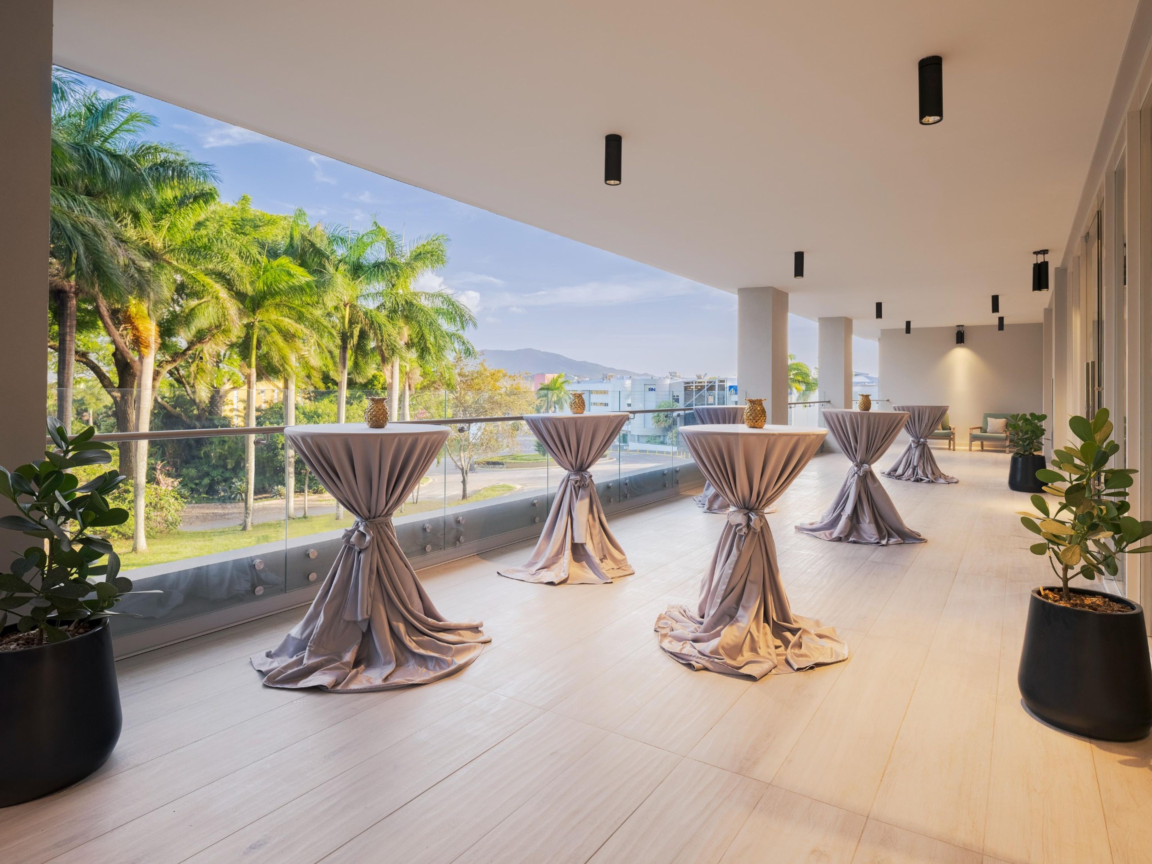 A spacious balcony with tables set for dining, overlooking a tropical landscape with palm trees and a clear sky.