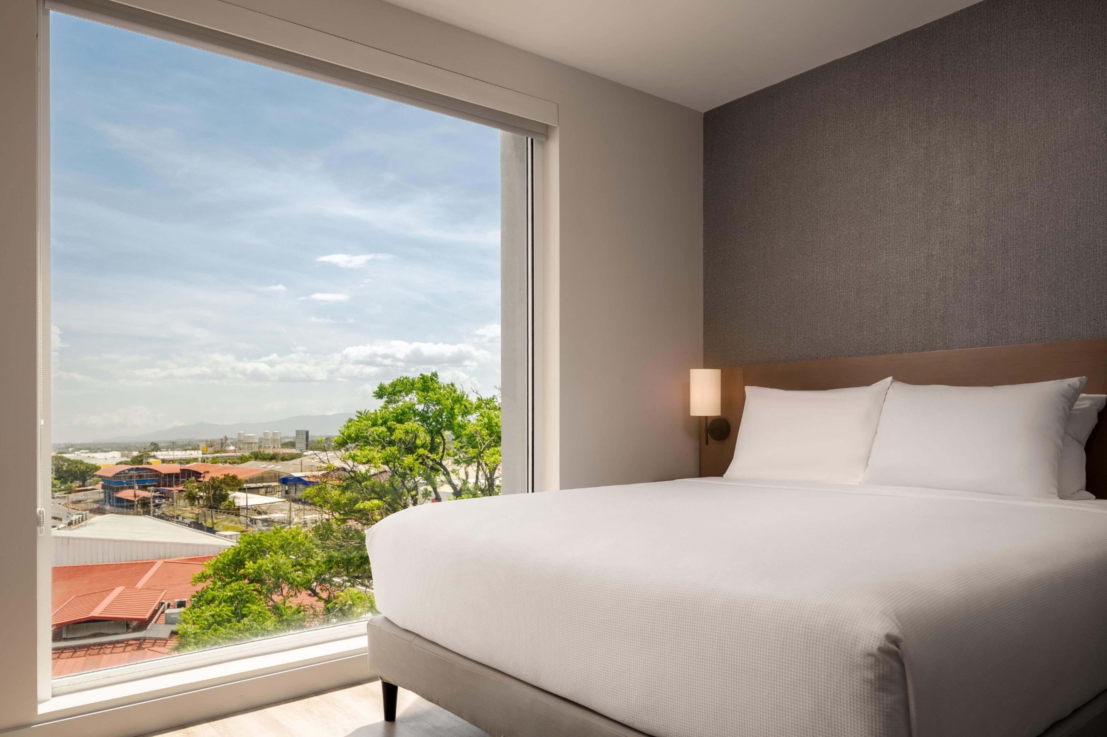 A hotel room with a large window, a bed, a nightstand, and a wall-mounted lamp.