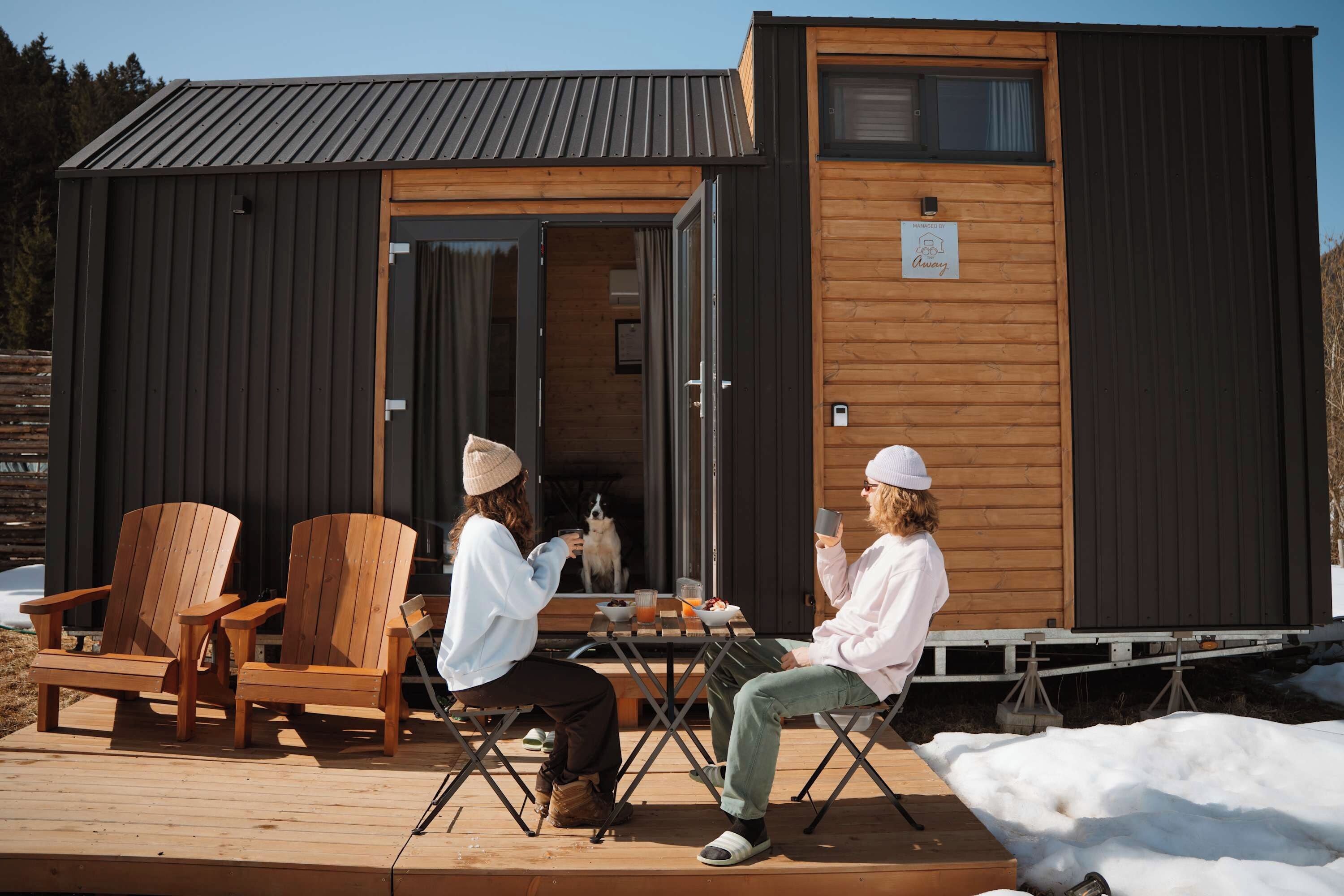 Eine Holzhütte mit einem Metalldach, eine Terrasse mit Holzstühlen und einem Tisch, an dem ein Hund und Futter stehen.