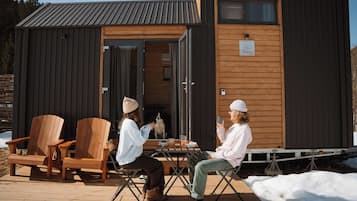 A wooden cabin with a metal roof, a deck with wooden chairs, and a table with a dog and food.