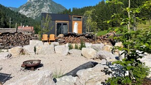 Una cabaña moderna con terraza de madera, chimenea de piedra y montañas al fondo.