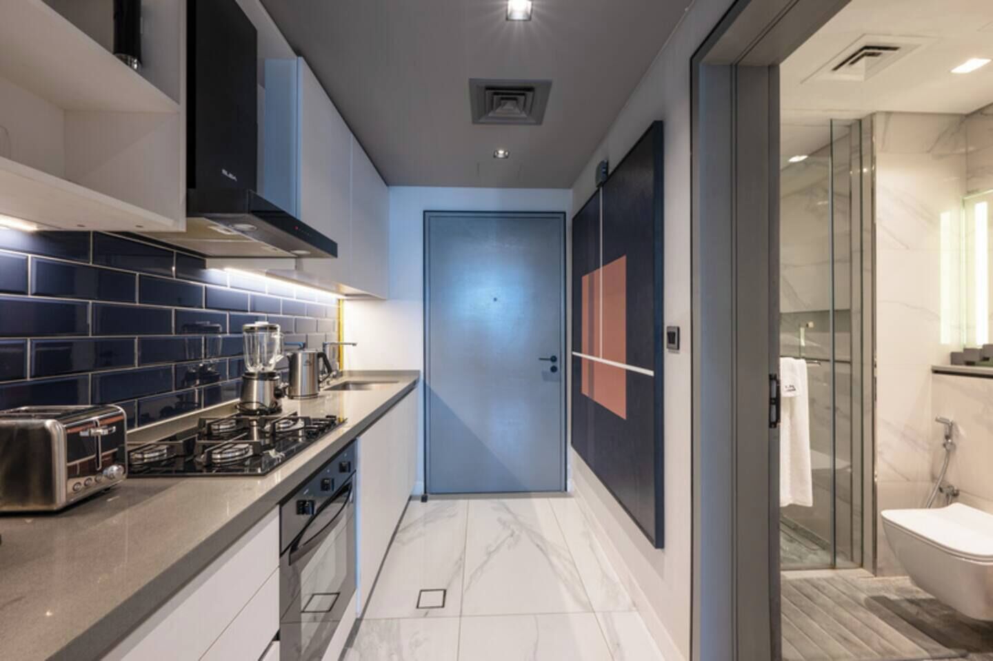 A modern kitchen with a gas stove, toaster, and coffee maker, leading to a bathroom with a glass shower and toilet.