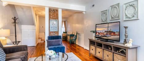 Amplia sala de estar con televisor grande, una mesa de centro y un sofá. Se aprecia una pared de ladrillo y una escalera que conduce a otra habitación.