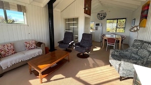 En rummelig stue med højt til loftet, en sofa, to lænestole, et sofabord og et spiseområde med bord og stole.