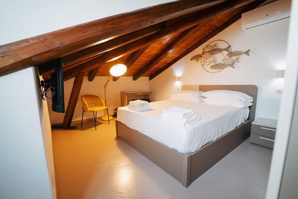 A bedroom with a sloped ceiling, a bed with white linens, a chair, and a wall-mounted light fixture.