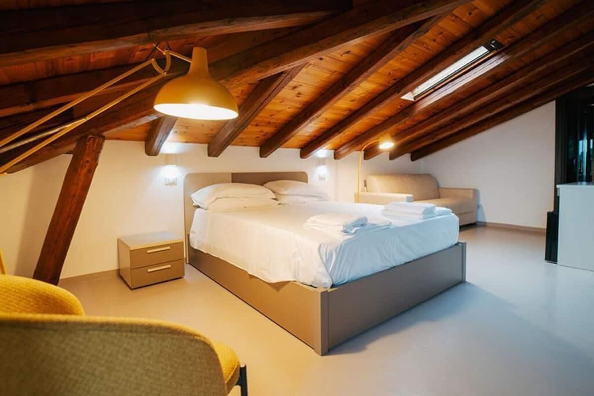 A modern bedroom with a wooden ceiling, a bed with white linens, a bedside table, and a chair.