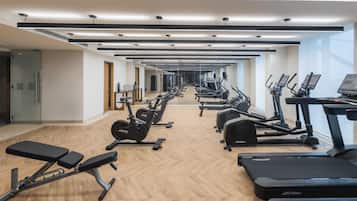 Uma academia com várias esteiras, elípticos e aparelhos de musculação, dispostos em um espaço amplo e bem iluminado, com piso de madeira e grandes janelas.