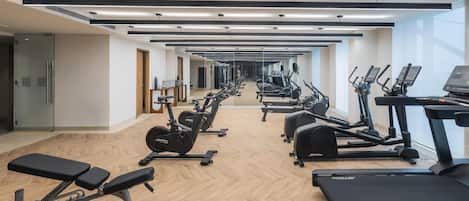 Uma academia com várias esteiras, elípticos e aparelhos de musculação, dispostos em um espaço amplo e bem iluminado, com piso de madeira e grandes janelas.