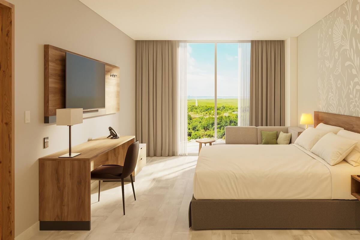 Ein modernes Hotelzimmer mit einem großen Bett, einem Schreibtisch, einem Sessel, einem Fernseher und Blick auf Grün durch das Fenster.