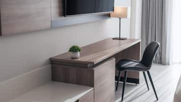A modern hotel room with a wooden desk, a black chair, a small potted plant, a flat-screen TV mounted on the wall, and a lamp on the desk.