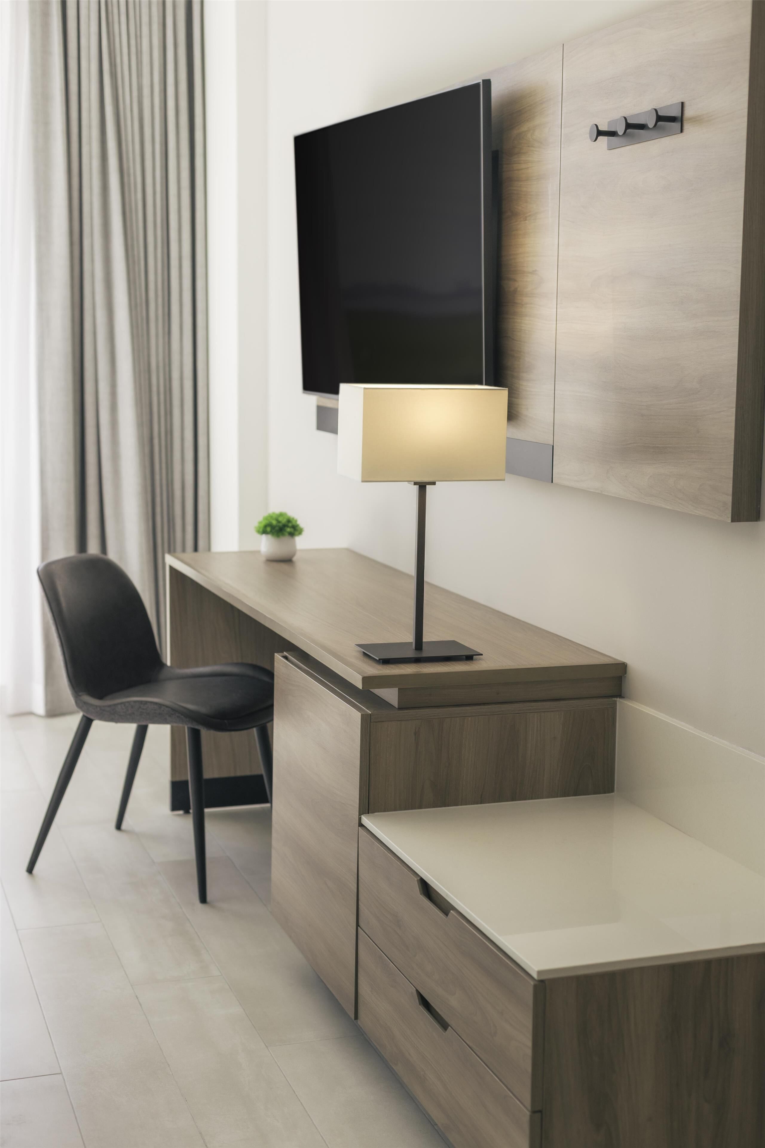 Una habitación de hotel moderna con un escritorio de madera, un televisor de pantalla plana montado en la pared, una lámpara de escritorio, una silla negra y una pequeña planta en maceta.