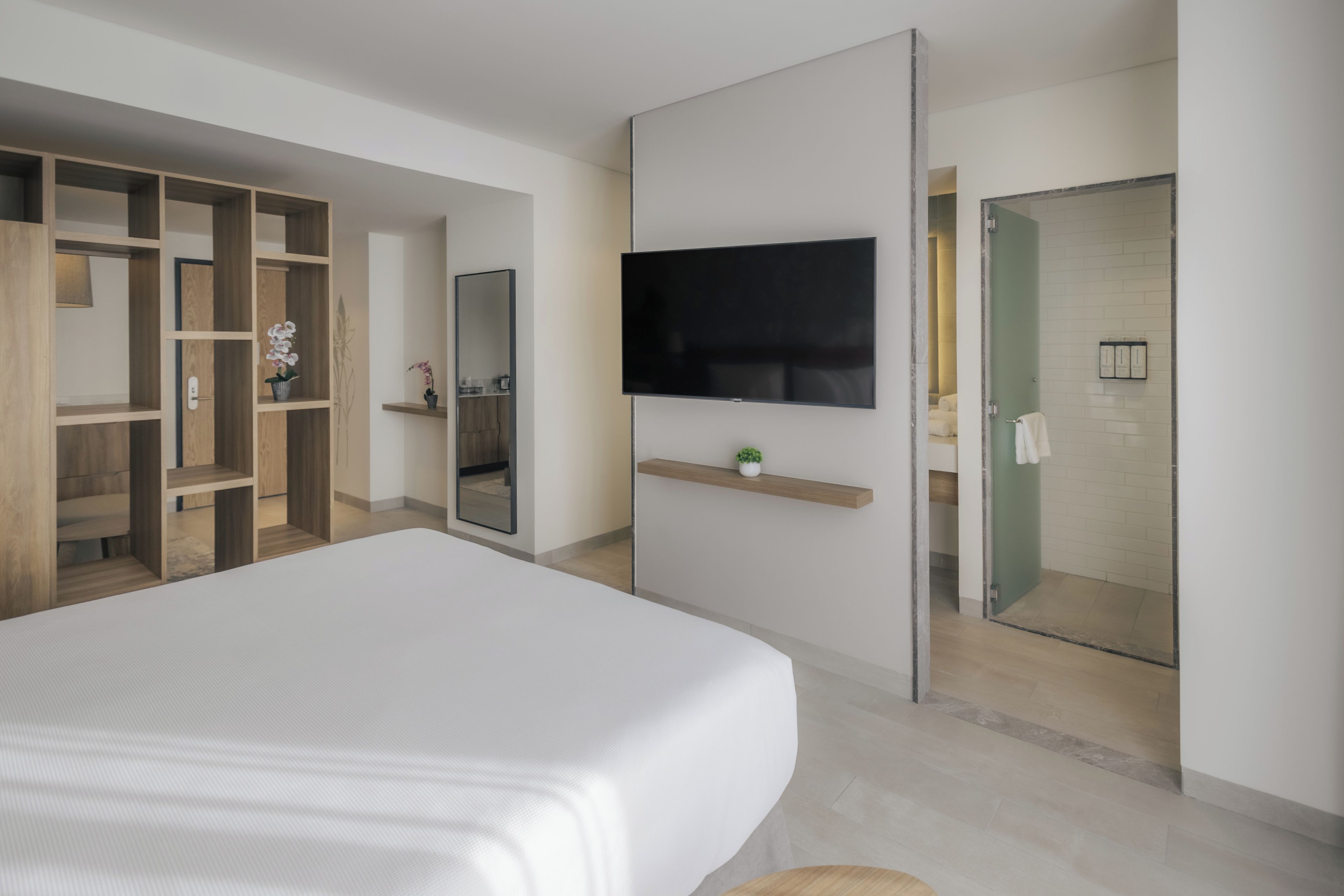 A modern bedroom with a large bed, a TV mounted on the wall, a wooden wardrobe, and a glass shower enclosure.