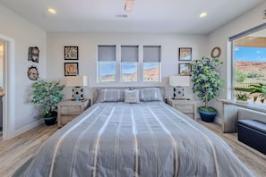 A bedroom with a large bed, two bedside tables with lamps, a window with blinds, and a view of a desert landscape.