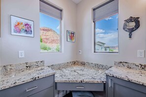 A corner kitchen with granite countertops, a window with a view of a mountain, and two framed artworks on the wall.