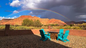 Een terras met twee turquoise Adirondack-stoelen en een tafel, met op de achtergrond rode rotsformaties en een regenboog.