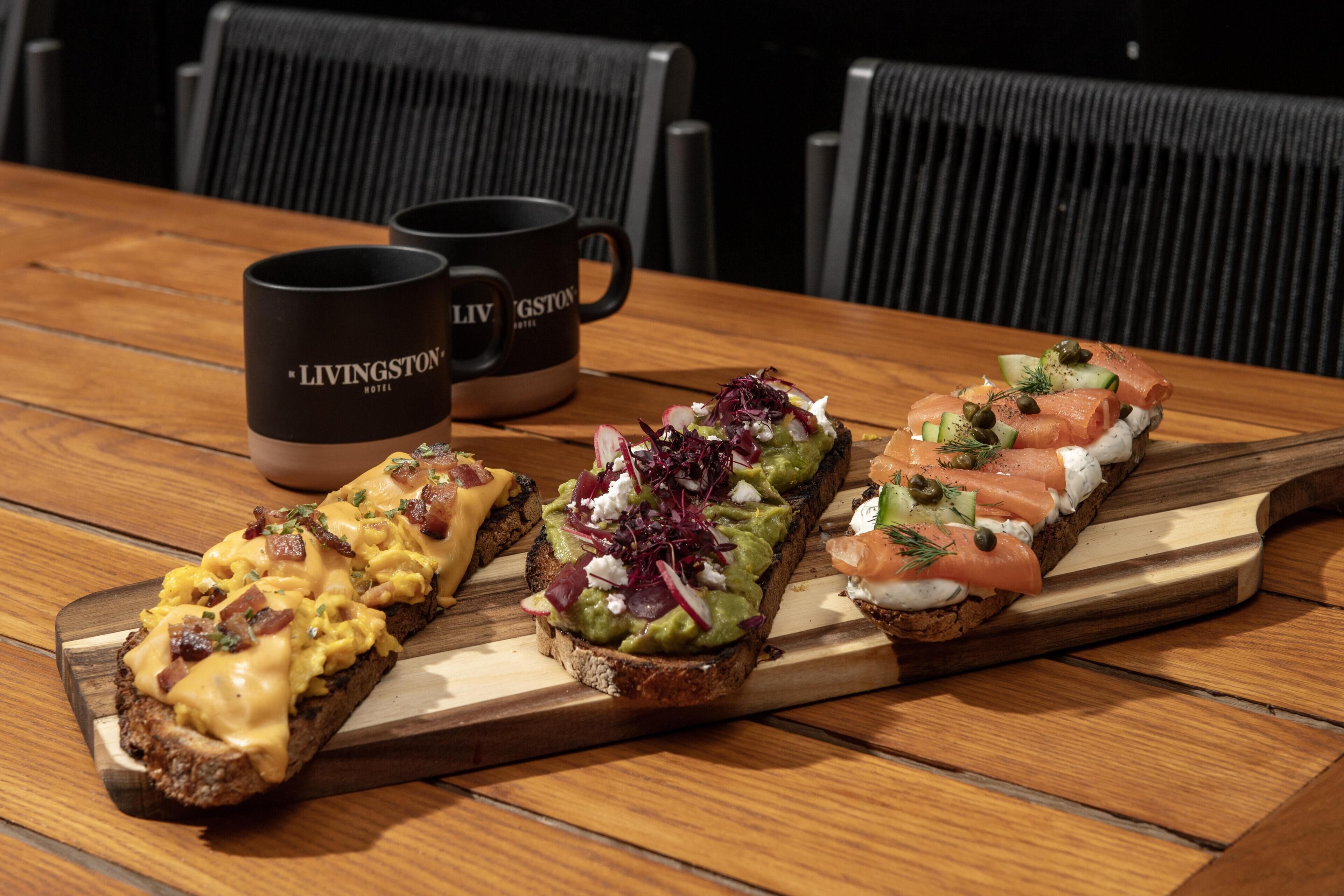 A wooden table with three open-faced sandwiches and two black mugs with 'LIVINGSTON HOTEL' text.