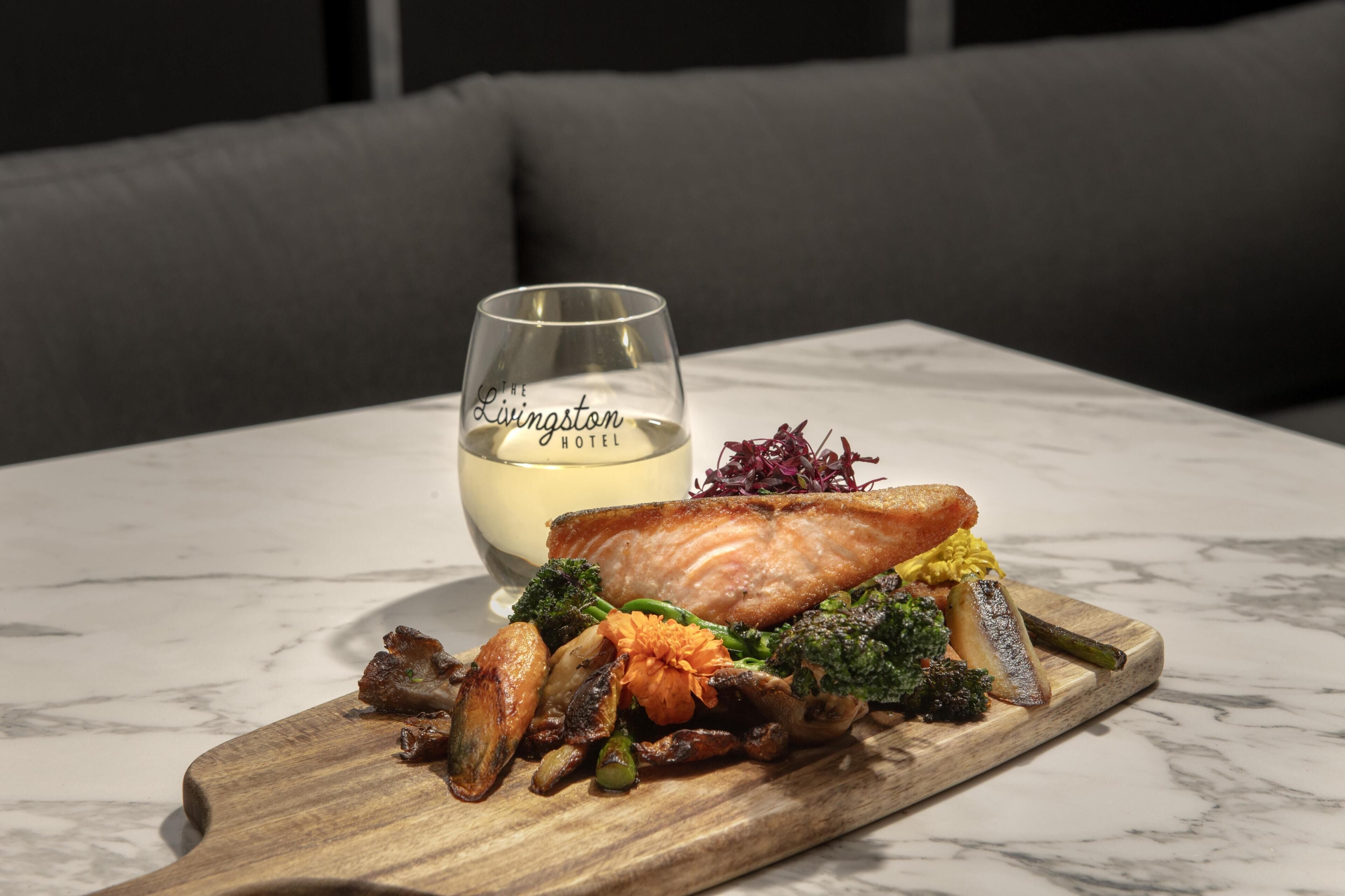 A wooden board with a meal, a glass of white wine, and a sofa in the background.