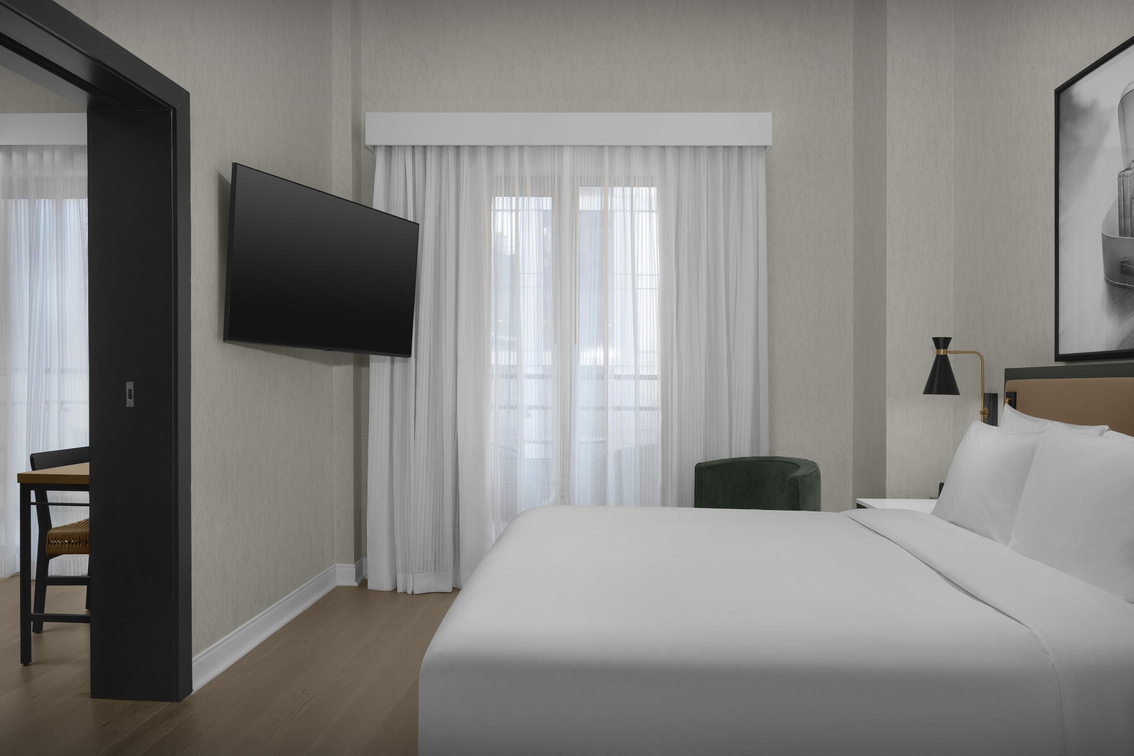A hotel room with a bed, a TV mounted on the wall, a desk with a chair, and a window with curtains.