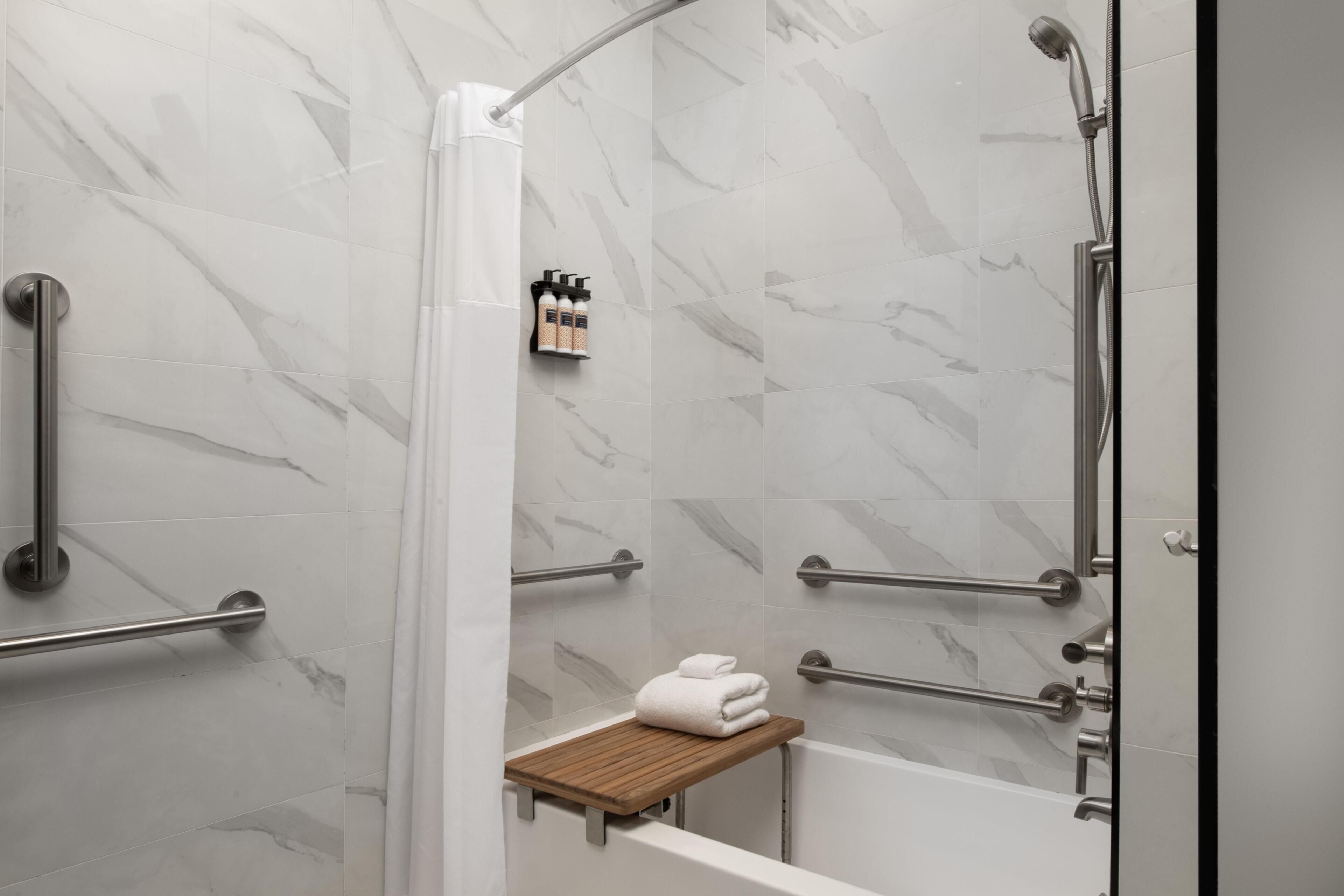 A modern bathroom with a shower, grab bars, a towel rack, and a wooden shelf.