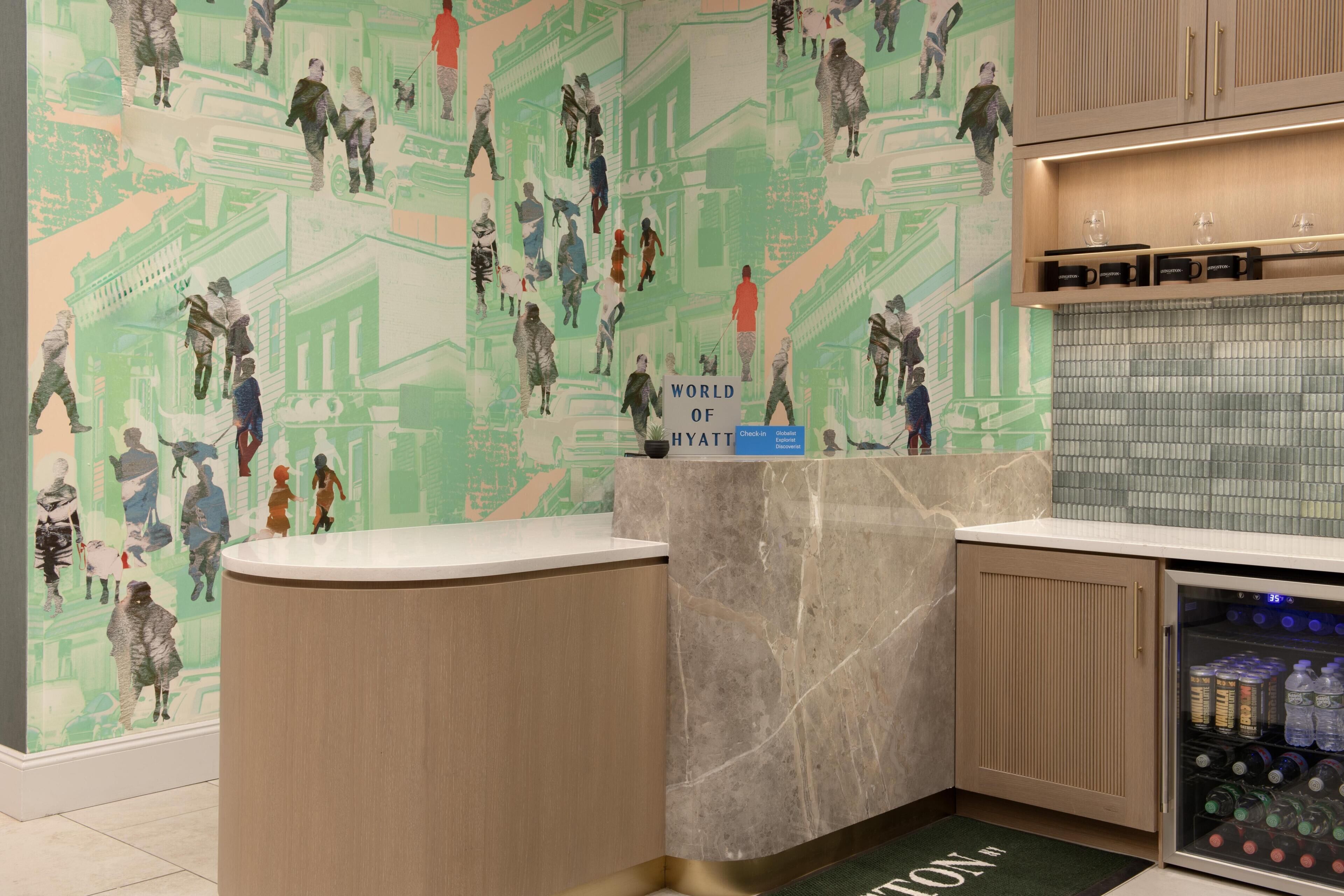 Zona de recepción de hotel con mostrador de mármol, una pared con un mural estampado en tonos verdes y una barra minibar con bebidas.