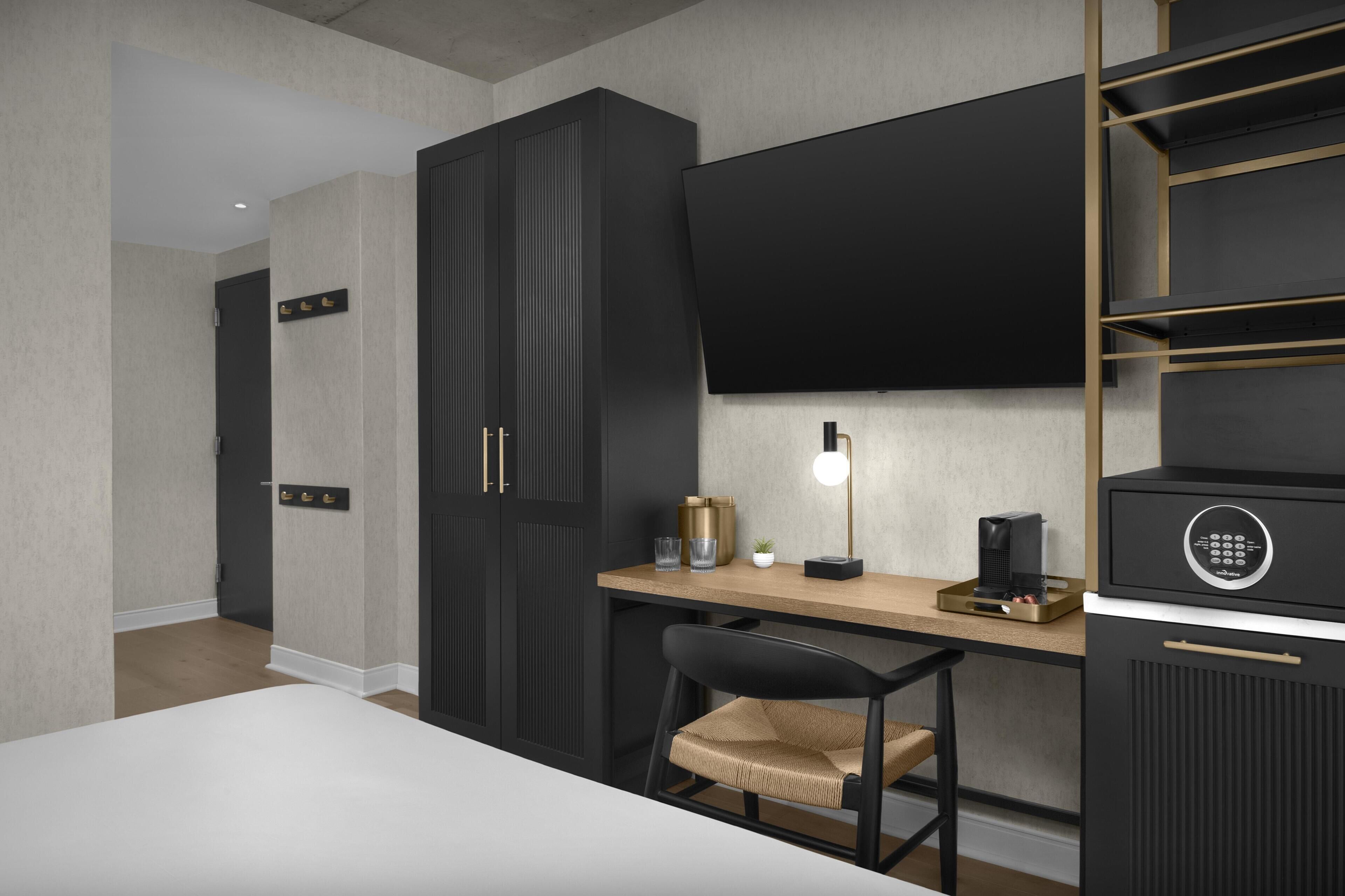 A modern hotel room with a bed, a desk with a chair, a television, and a minibar.