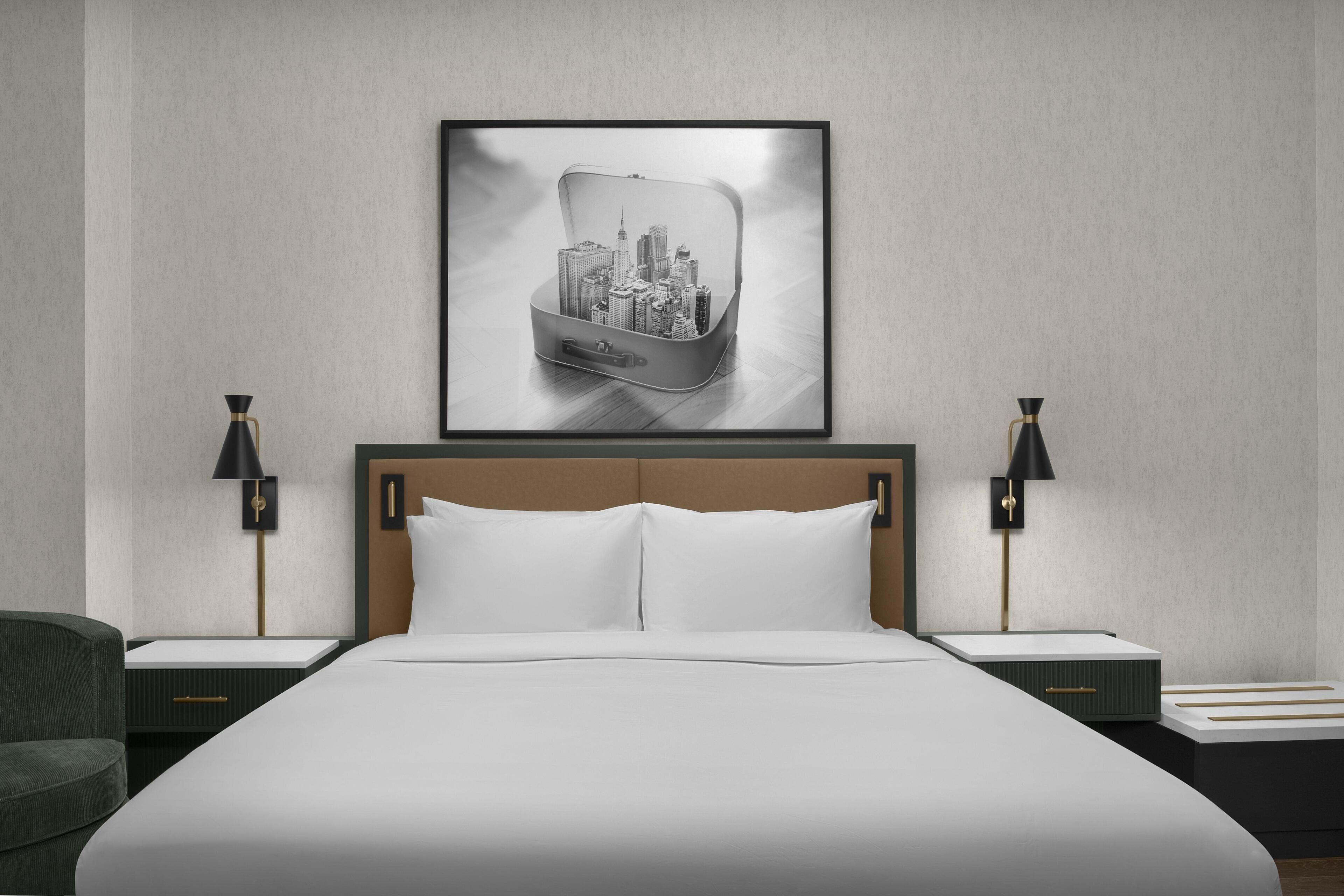 A hotel room with a large bed, two bedside tables, a headboard, and a framed cityscape picture on the wall.