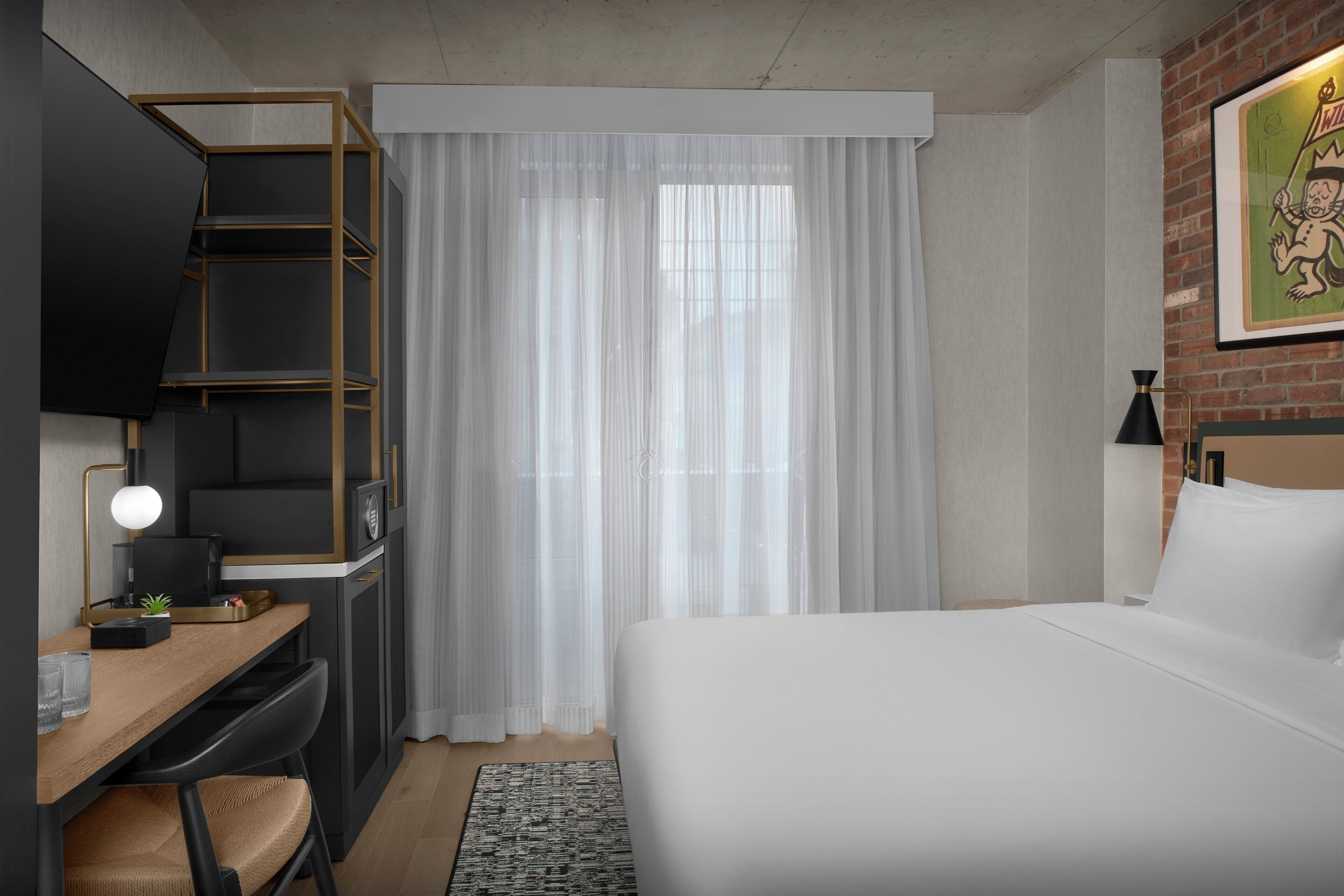 A hotel room with a bed, desk, chair, and a large window with sheer curtains.