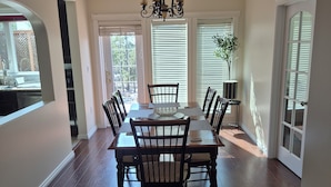 A dining area with a wooden table and chairs, a chandelier, and a view of the outdoors through large windows.