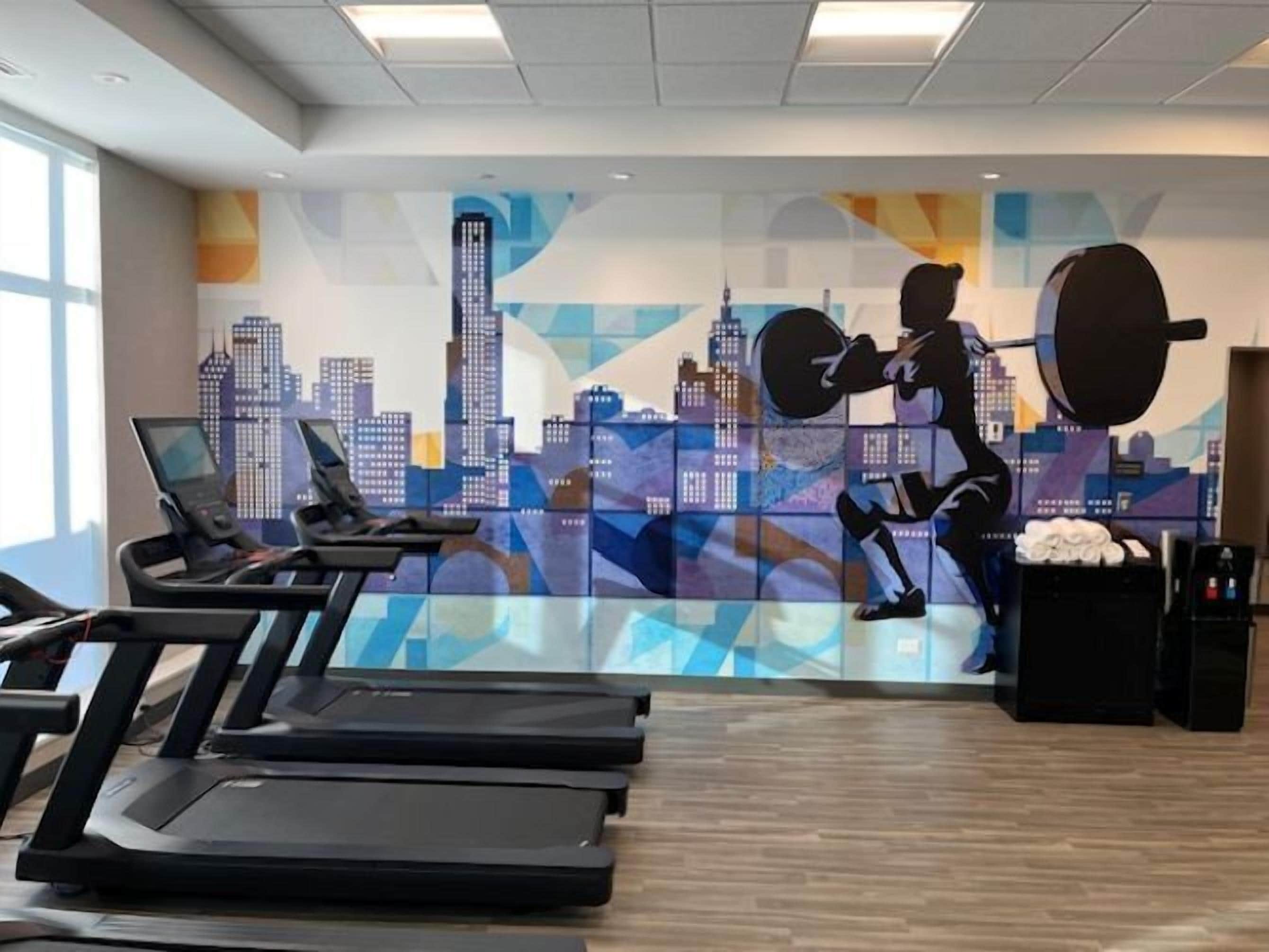 Un gimnasio con cintas de correr, zona de pesas y un mural con vistas a la ciudad.